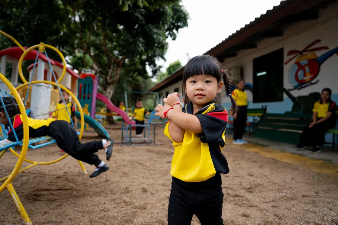 A young girl in a yellow school uniform stands in a playground with her arms crossed, while other children play on climbing equipment and adults sit near a school building behind her.