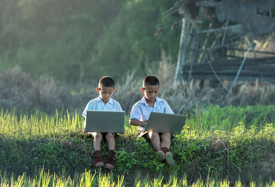 Two boys in student uniform are sitting next to each other on a grass field, using laptops.