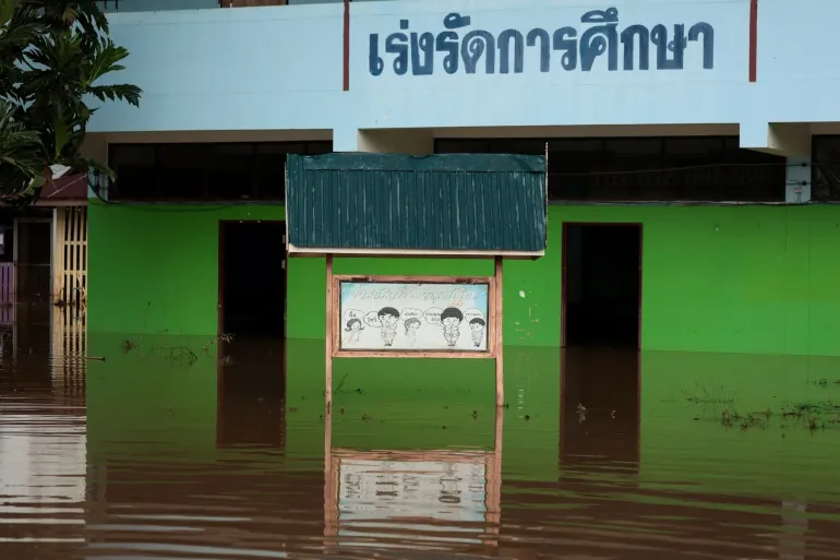 Floodwater surrounds the ground floor of a school building, reaching the doorways and lower green walls, with a notice board partly submerged in front.