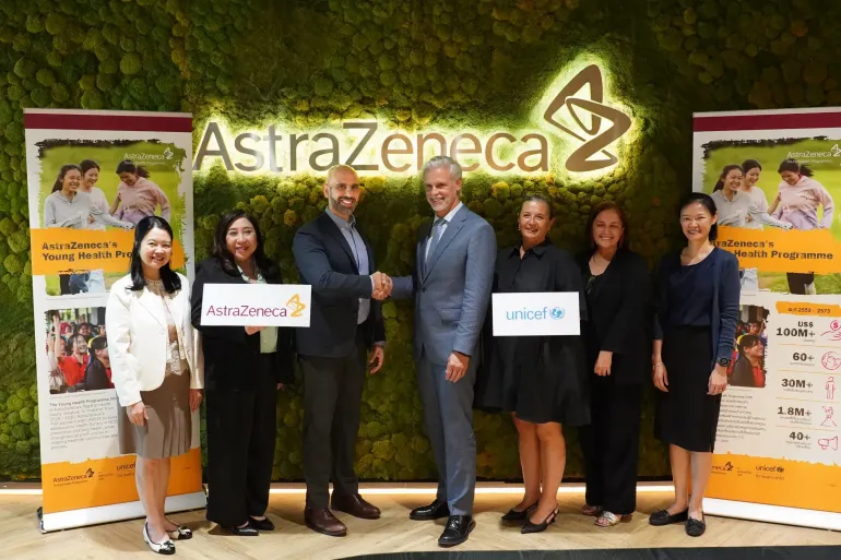 Seven adults pose at an AstraZeneca office, with two men shaking hands at centre and participants holding AstraZeneca and UNICEF signs, flanked by Young Health Programme banners.
