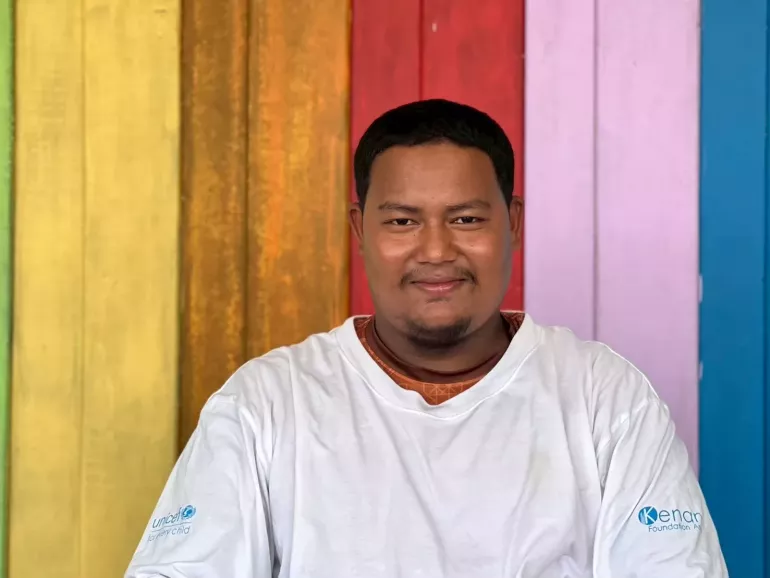 A man in a white shirt with UNICEF and Kenan Foundation Asia logos, smiling in front of a colorful wooden wall with vertical panels in yellow, red, purple, and blue.