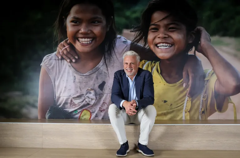 Ken Legins, Representative for UNICEF thailand, in a suit sits smiling in front of a large photo of two children laughing with their arms around each other.