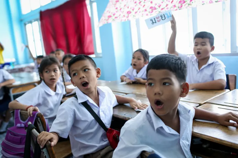 Students in white uniforms participate in a classroom activity, with several children speaking and one boy holding up a card with Thai letters.