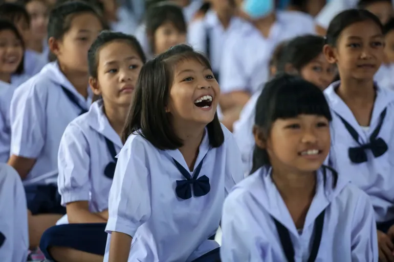 A group of schoolchildren in white uniforms sit together, with a girl in the center laughing.