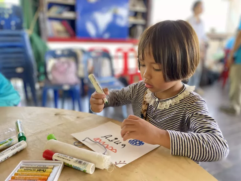 A young girl with short hair and wearing a striped shirt concentrates as she draws on a piece of paper at a round table. Crayons and markers are scattered on the table. The background shows a casual indoor setting with plastic chairs and blurred figures.