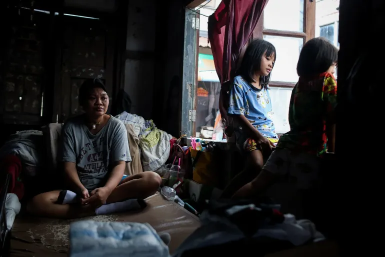 A woman sits on a mattress inside a dimly lit home while two children sit by a bright window beside her.