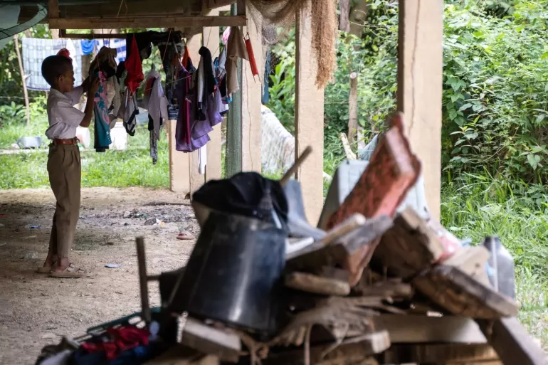 A young boy wearing a white shirt and brown pants is hanging clothes on a clothesline under a raised house. Various garments are draped over the line, and the area appears rural, with scattered items and greenery in the background. The foreground shows a pile of old materials and tools, adding to the rustic atmosphere.
