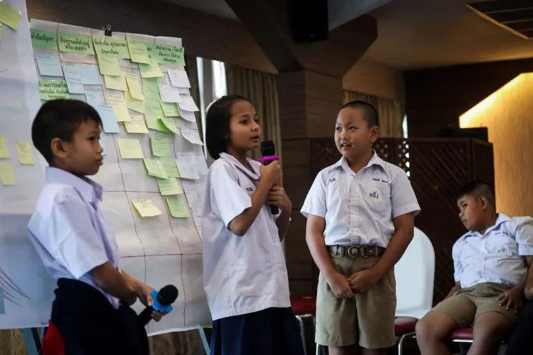 Group of students in school uniforms presenting in front of a board covered with sticky notes during workshop activity.