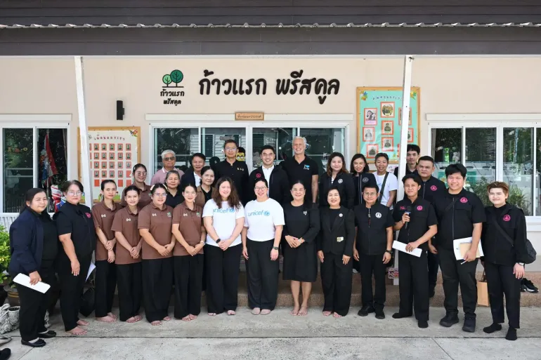 กลุ่มผู้ใหญ่ รวมถึงผู้ที่สวมเสื้อยูนิเซฟสองคน ยืนถ่ายภาพร่วมกันบริเวณทางเข้า &ldquo;ก้าวแรก พรีสคูล&rdquo;