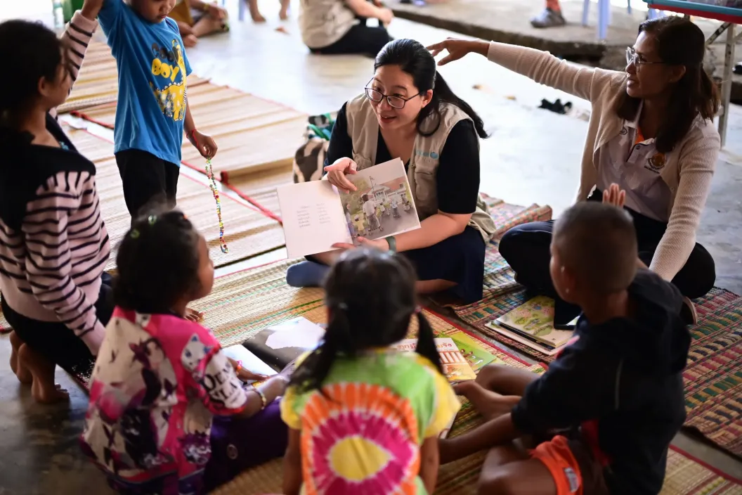 An UNICEF staff reads a picture book to a small group of children sitting on mats, while another adult gestures to engage them during a group activity.