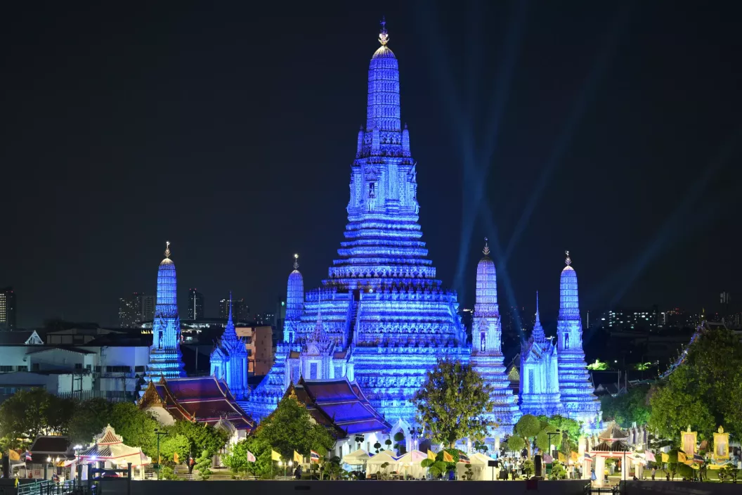 พระปรางค์วัดอรุณราชวรารามในกรุงเทพฯ ส่องแสงไฟสีน้ำเงินยามค่ำคืน พระปรางค์ใหญ่และพระปรางค์เล็กรอบข้างเปล่งประกายสว่างงดงามตัดกับท้องฟ้ามืด ด้านล่างมีต้นไม้และสถาปัตยกรรมไทยแบบดั้งเดิม พร้อมแสงไฟจากตัวเมืองที่มองเห็นอยู่ไกล ๆ