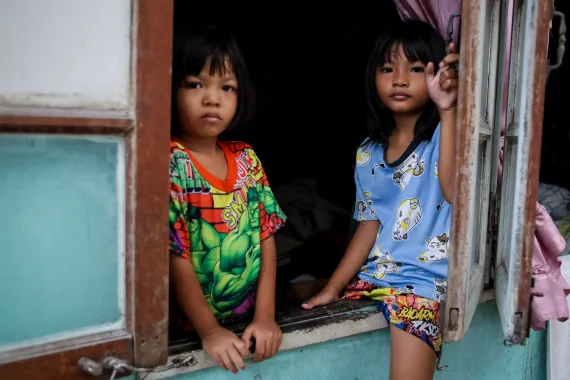 Two young children look out from an open wooden window; one stands in a colourful shirt while the other sits on the sill in a blue cat-print shirt.