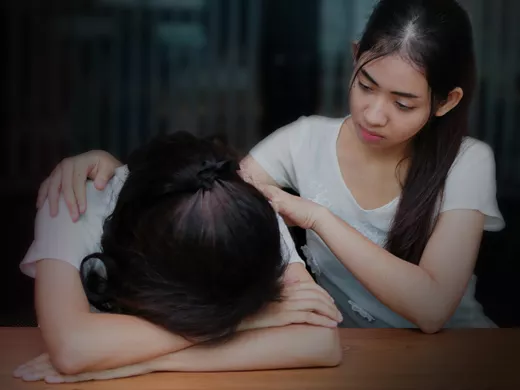 An emotionally charged scene in a dimly lit room: one individual rests her head on a wooden table, seemingly upset. Her companion is offering comfort.
