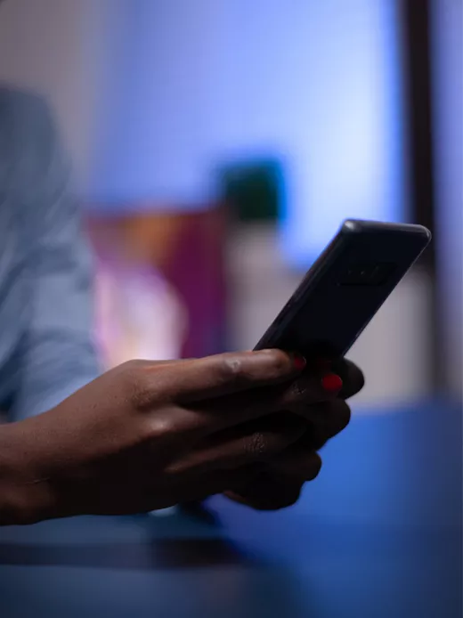 A hand holds a smartphone, illuminated in the soft glow of a dimly lit room. The focus is on the hand and phone, while the background is blurred because it's out of focus.