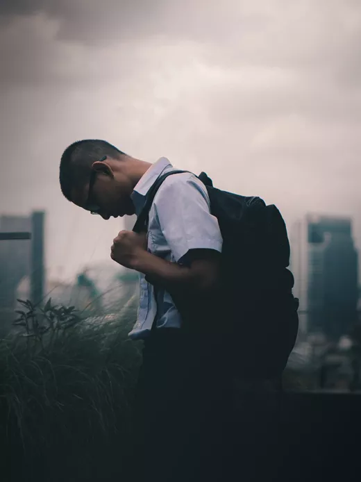 A male student in a white shirt and black pants, carrying a black backpack, is walking along the street. The city skyline looms in the background. The sky above the city is cloudy, adding a dramatic touch to the scene.