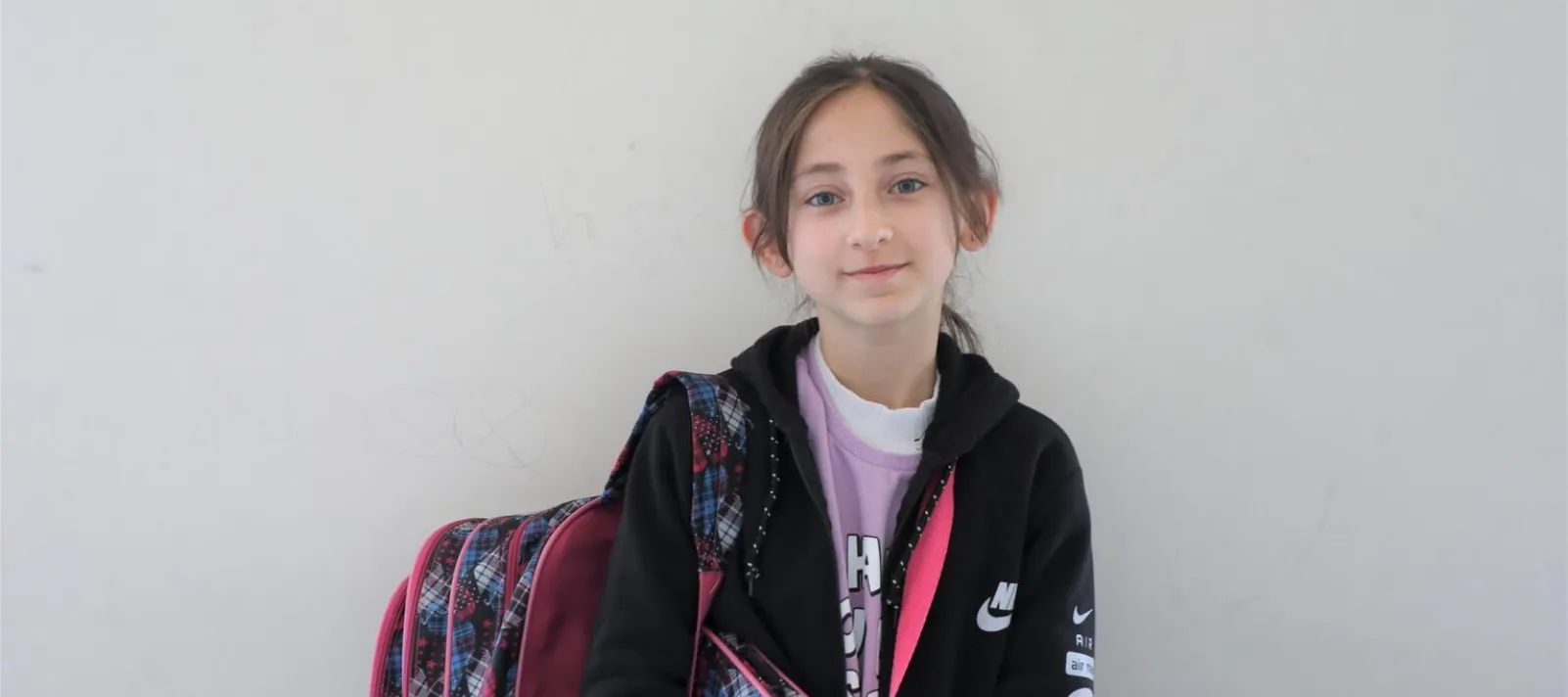 A young girl smiling to the camera holding her backpack. 