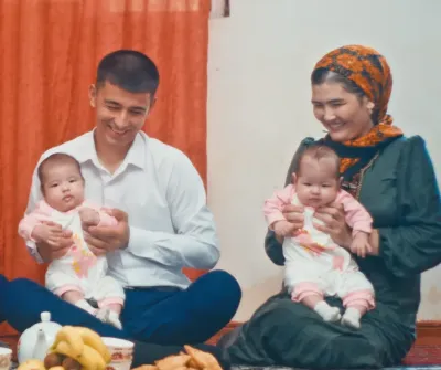 A woman and a man sit on a carpet, holding two babies and two young children, with fruit, pastries and tea laid out in front of them.