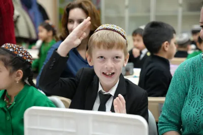 A young boy wearing a traditional skullcap smiling brightly and raising his hand during a lesson in an inclusive cl
