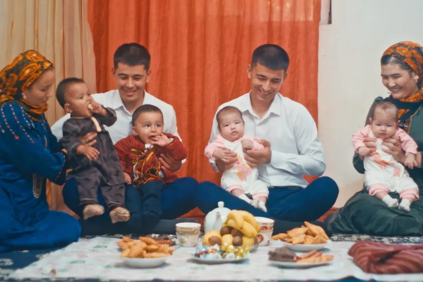 Two women and two men sit on a carpet, holding two babies and two young children, with fruit, pastries and tea laid out in front of them.