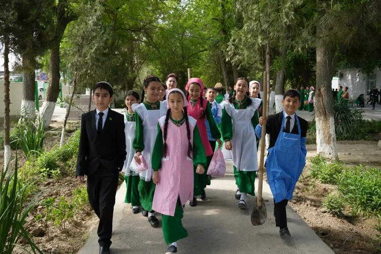 Schoolchildren in Turkmenistan walk along a tree-lined school path during an environmental activity, with one student carrying a shovel and others wearing aprons as they take part in keeping their school grounds clean and green.