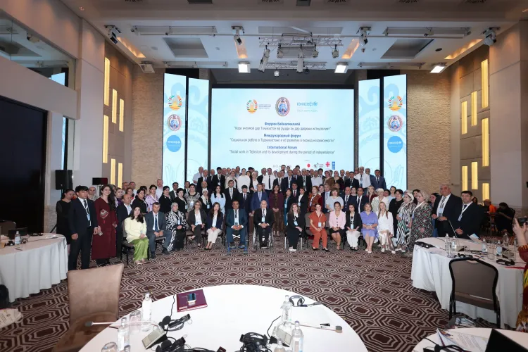 A large group of delegates, speakers, and organizers posing together on stage at the International Forum on Social Work in Dushanbe. A large digital backdrop displays event branding and titles in Tajik, Russian, and English.