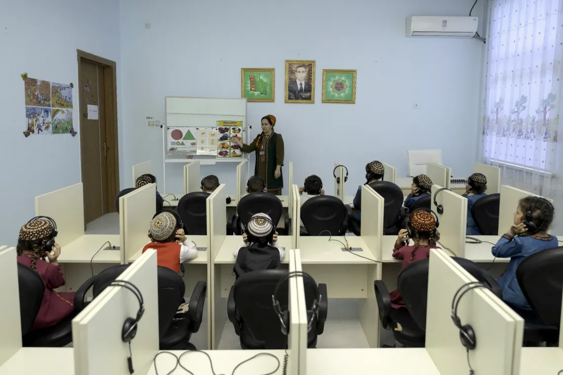 A teacher leads a lesson for children in a classroom equipped with individual computer stations and headphones.