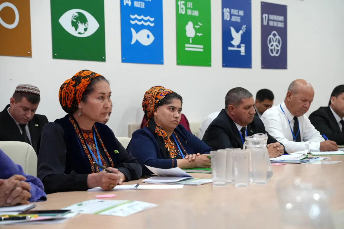 Participants seated at a conference table taking notes during a meeting, with Sustainable Development Goals (SDGs) posters displayed on the wall behind them.