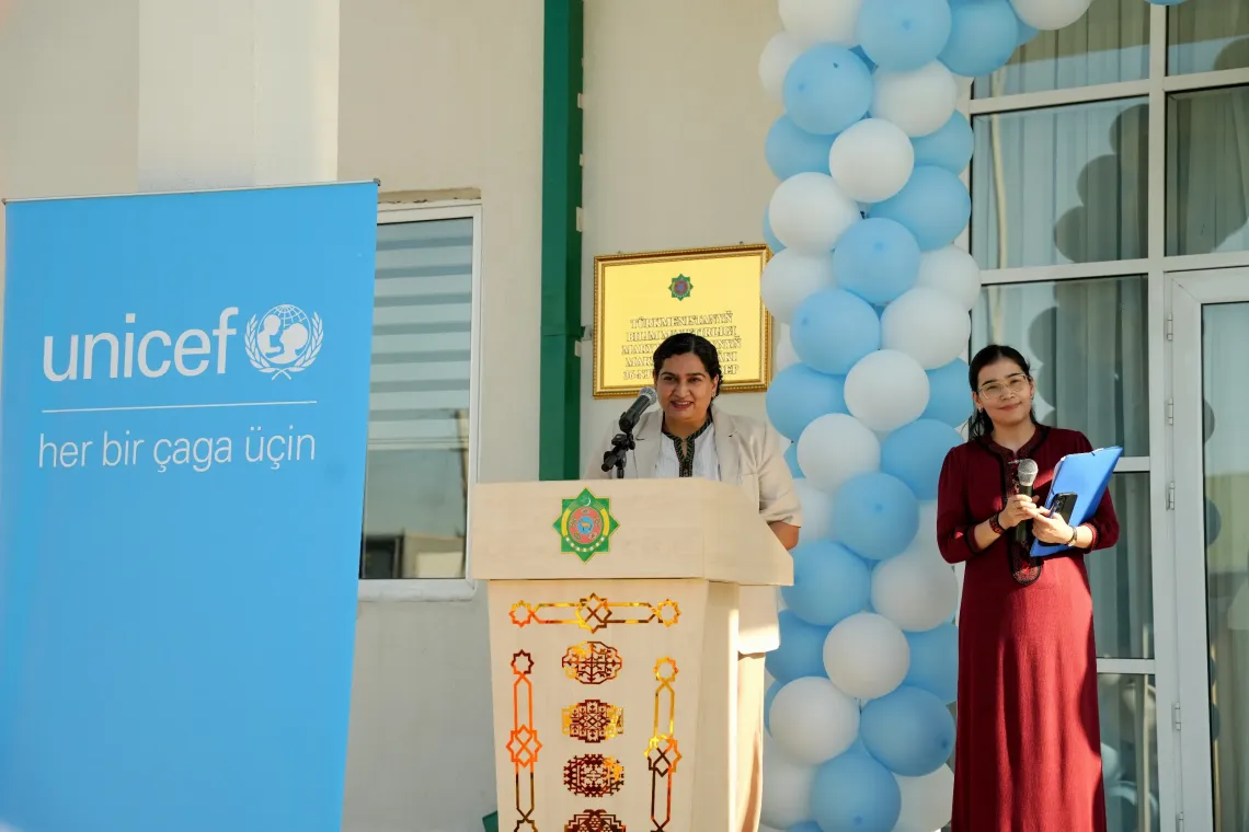 UNICEF Representative Jalpa Ratna speaking at a podium next to a blue UNICEF banner and a balloon arch during the opening ceremony.