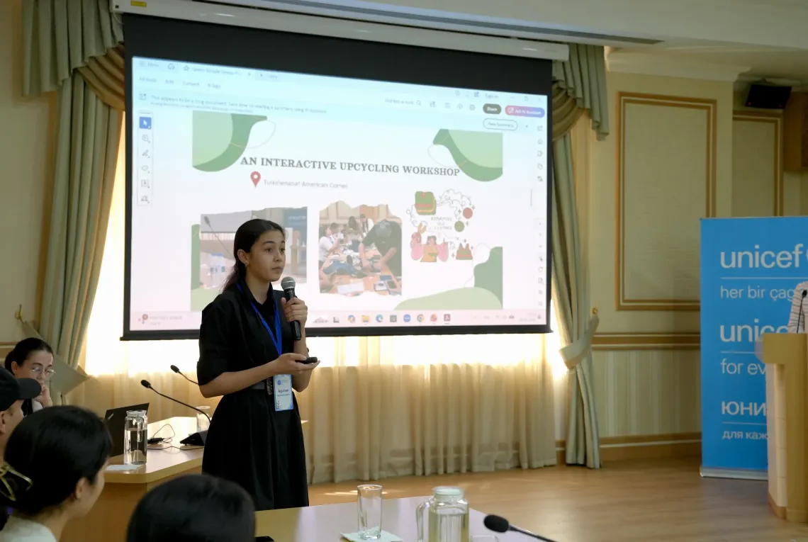 A young woman holds a microphone and presents a slide titled "An Interactive Upcycling Workshop" to the audience.