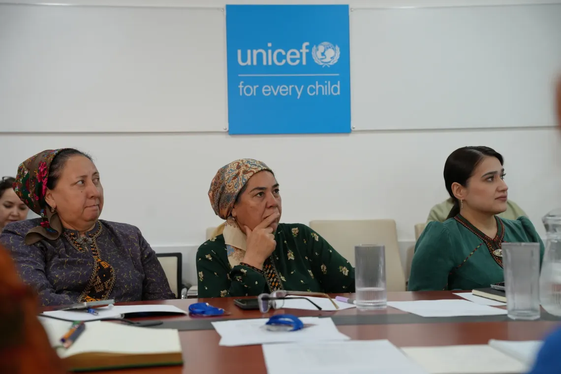 Three women sitting at a conference table, listening attentively during a meeting with a blue UNICEF "for every child" sign mounted on the wall behind them.