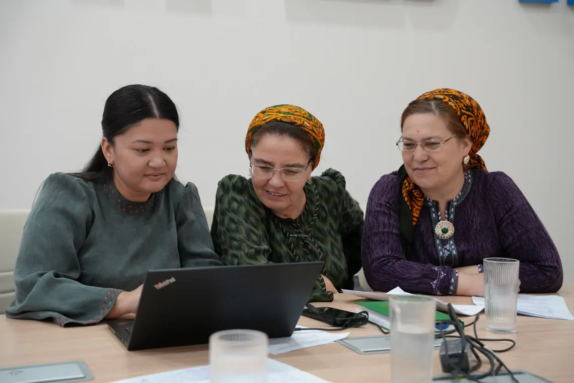 Three women collaborating and looking closely at a laptop screen together during a working session at a conference table.