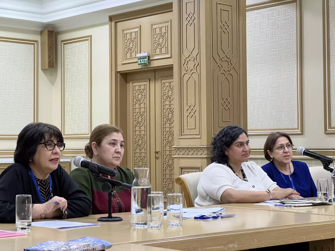 Four women at a conference table, two speaking into microphones, in a formal setting.