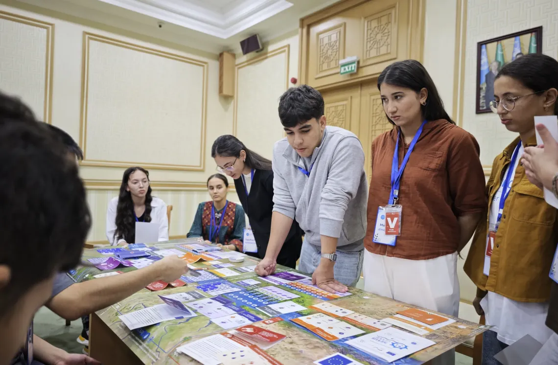 A group of young participants gathered around a table engaged in an interactive simulation board game featuring maps, task cards, and learning materials.