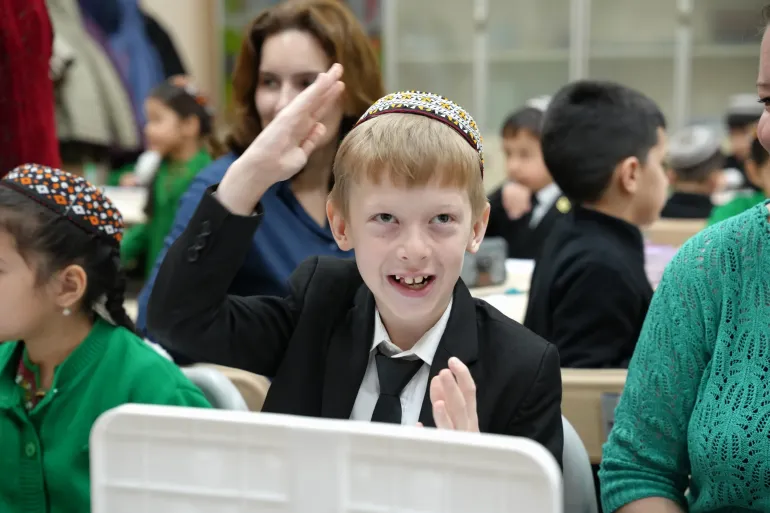 A young boy wearing a traditional skullcap smiling brightly and raising his hand during a lesson in an inclusive cl