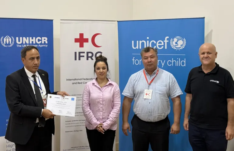 Four workshop organizers and participants standing in front of UNHCR, IFRC, and UNICEF banner stands, holding a newly awarded certificate of participation.