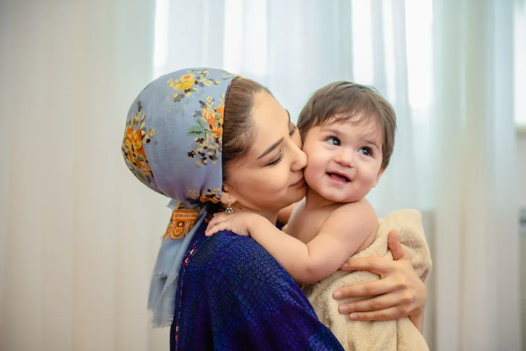 Woman in a floral headscarf kisses a smiling baby in her arms indoors.