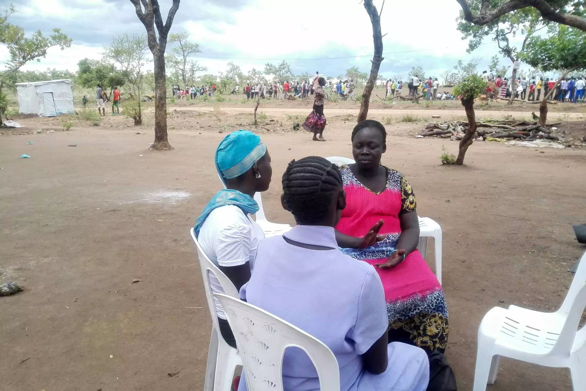 Two adolescent girls and one woman sit on white plastic chairs in the shade of a tree