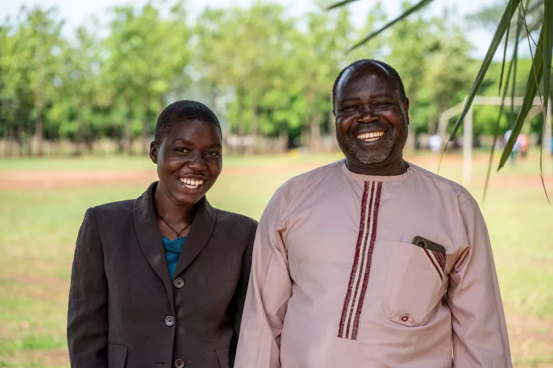 Beatrice Aber and her father, Daniel Uhuru, pose under a tree at Palabek Secondary School in Lamwo District after receiving their CashPlus funds. &ldquo;We lost everything, including her mother, when we fled,&rdquo; Daniel says. &ldquo;But I didn&rsquo;t want her to lose school too.&rdquo;