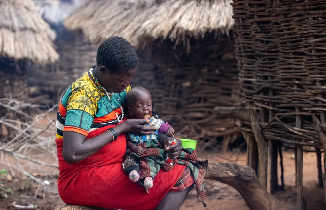Lokol Regina feeds one of her twins with Ready-to-Use Therapeutic Food (RUTF) at their home in Leterwa village, Kaabong. The high-calorie, nutrient-dense paste is the cornerstone of Severe Acute Malnutrition treatment for children without medical complications, and can be administered at home, allowing Regina to keep her children in treatment without leaving her household behind.