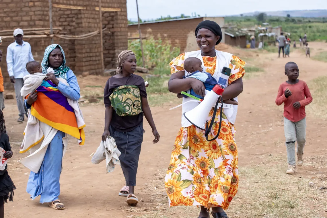 Uwase Christine, a Village Health Team (VHT) member, walks with a child and a group of mothers to a nutrition outreach session in Nakivale Refugee Settlement, Isingiro District. UNICEF-trained VHTs like Christine move through the settlement to mobilise families, screen children for malnutrition using MUAC tapes, and refer those at risk to treatment and feeding support. The work is supported by the Uganda Humanitarian Fund through UNICEF.