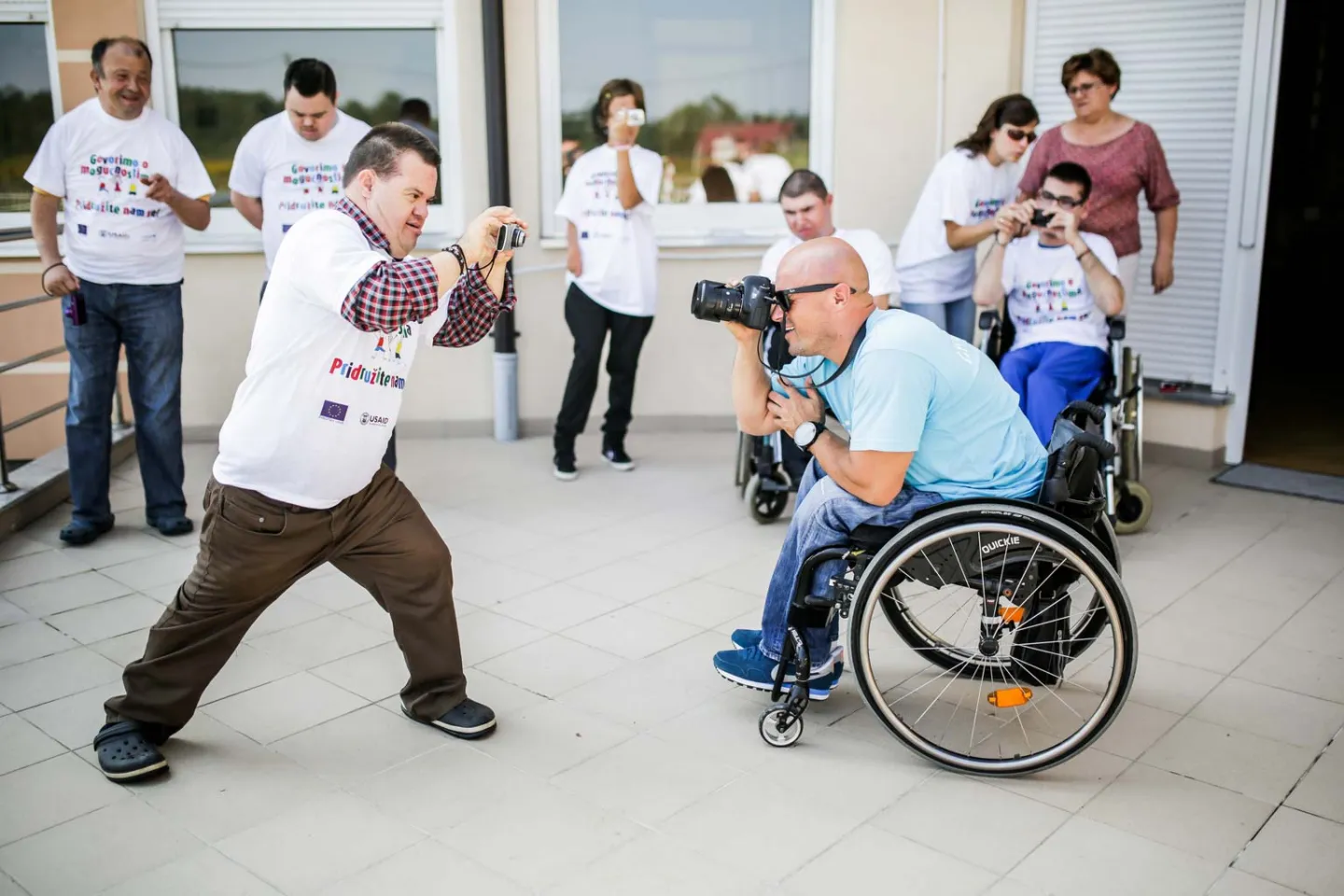 Two young men with disabilities playfully point cameras at each other.