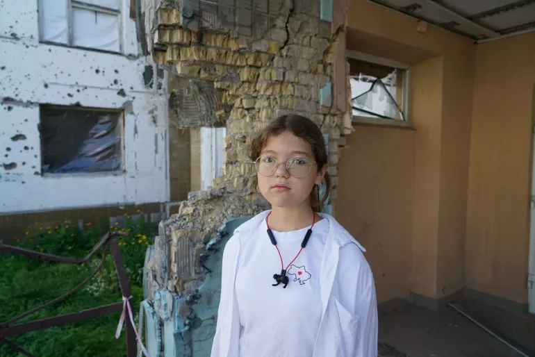 Дівчинка стоїіть біля стіни школи, в якій є сліди від снарядів та куль