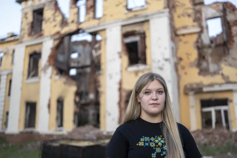 Дівчина із довгим світлим волоссям стоїть перед зруйнованою жовтою стіною школи