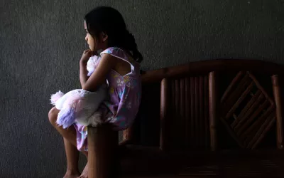 Una ni&ntilde;a sentada en el borde de un banco de madera mira hacia adelante mientras agarra un peluche. La ni&ntilde;a se ve de perfil.