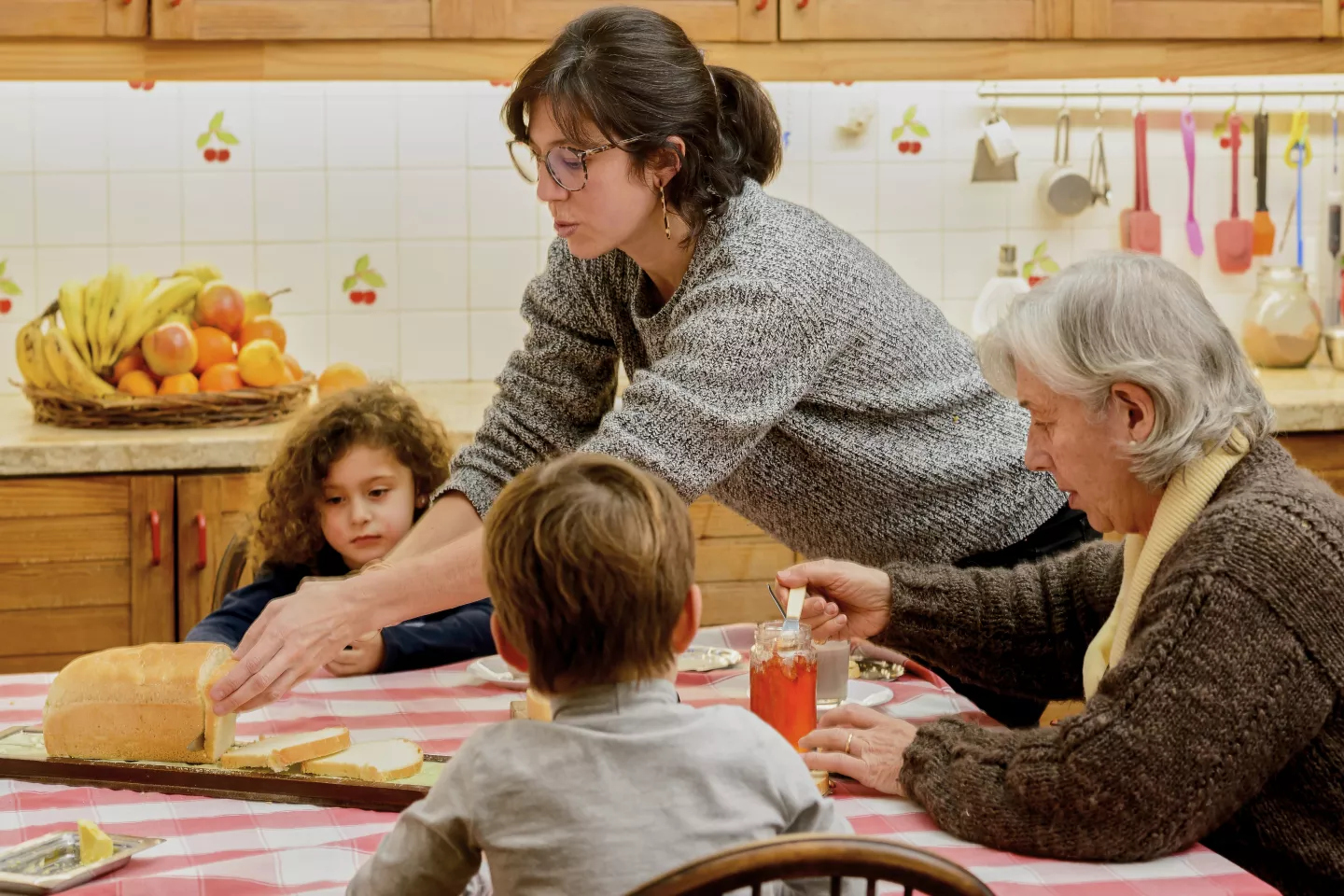 Mujer cortando pan sobre mesa, donde est&aacute;n sentados un ni&ntilde;o, una ni&ntilde;a y una se&ntilde;ora mayor.