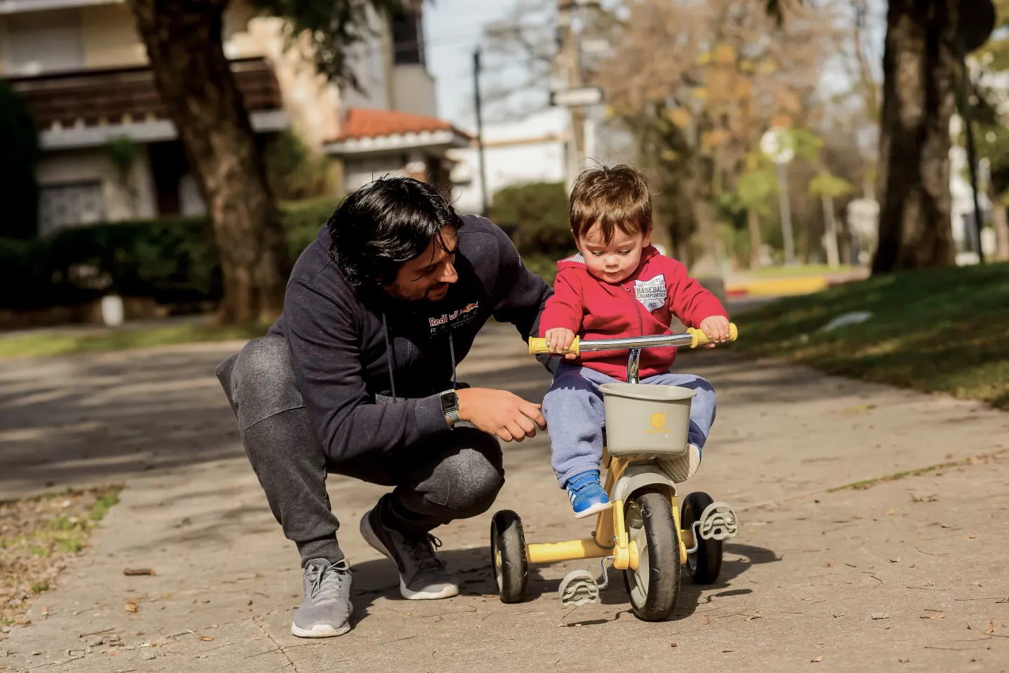 Padre acompa&ntilde;a a su hijo que anda en triciclo, en una plaza.