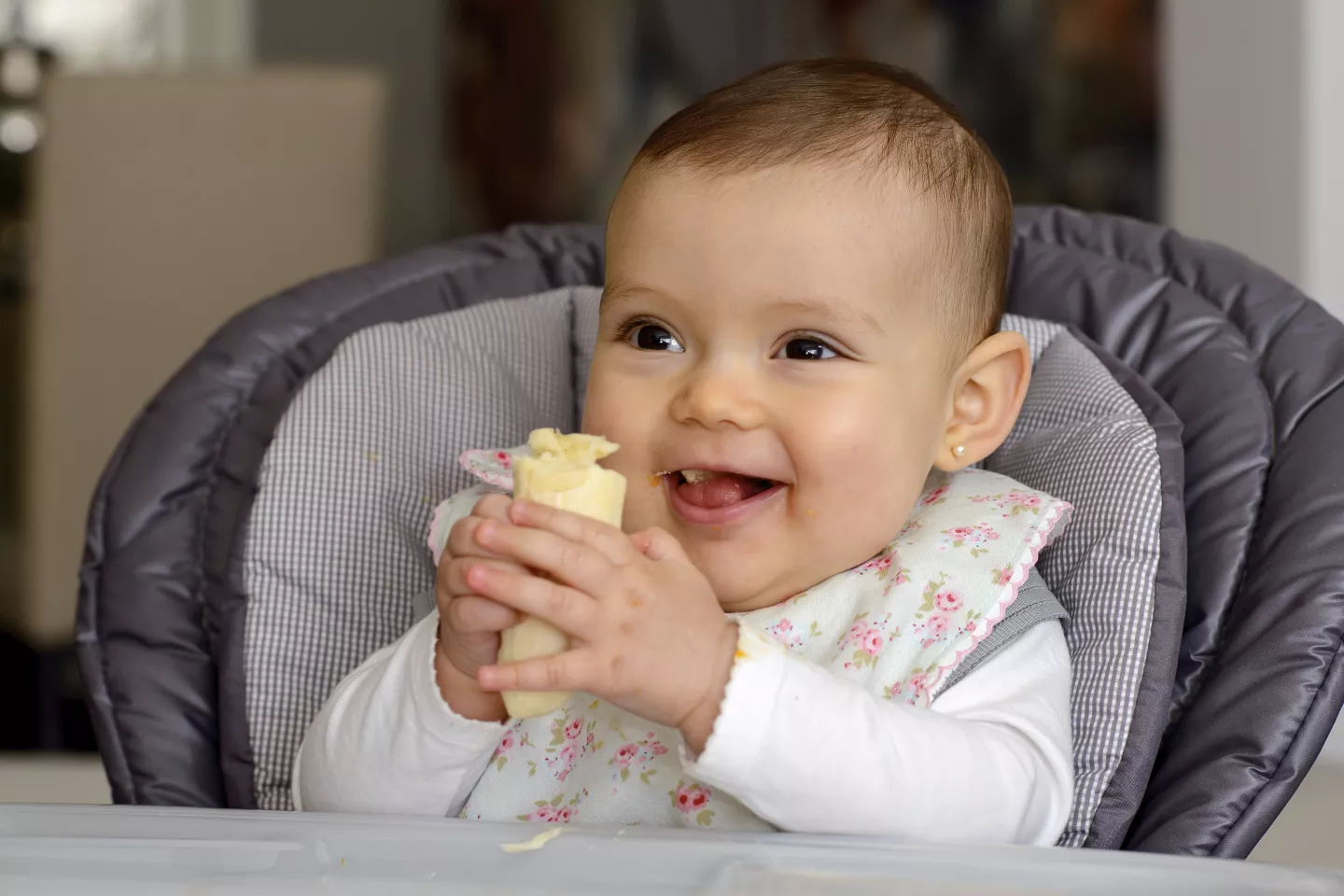 Beb&eacute;, sentada en sillita de comer, agarrando y comiendo media banana.