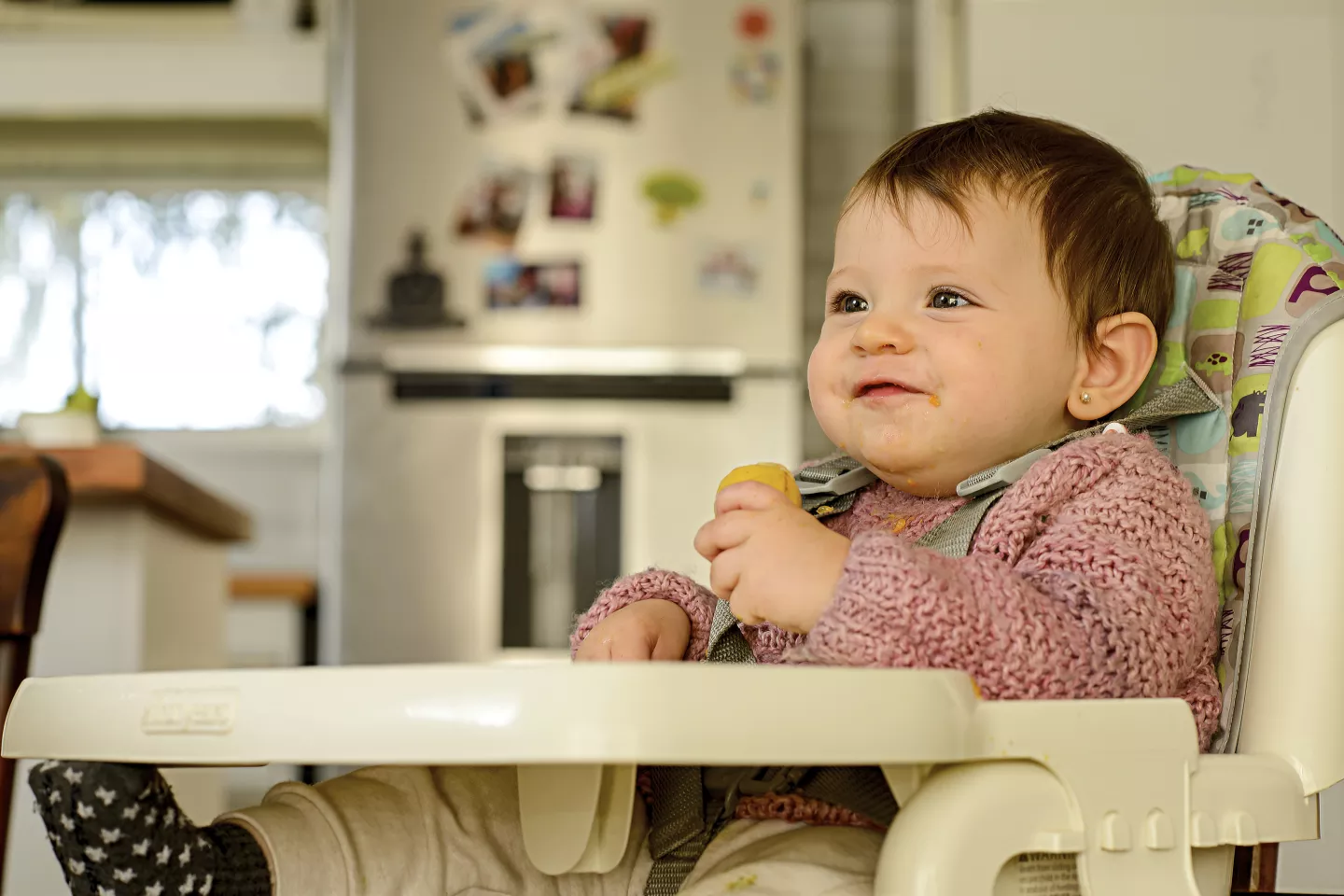 Beb&eacute; de unos 8 meses, sentada en sillita de comer, agarrando una fruta con la mano.