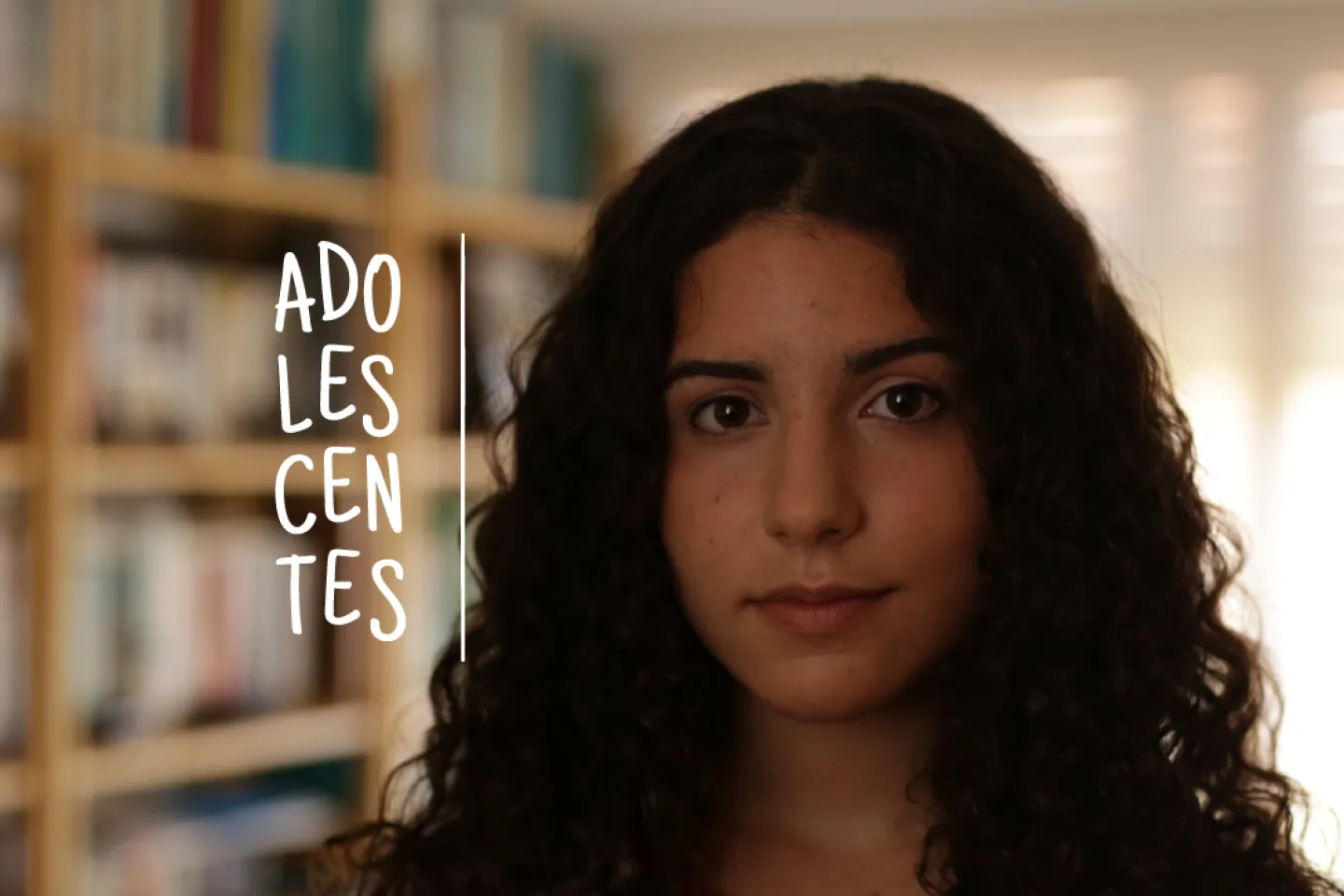 Retrato de adolescente mujer, en una habitaci&oacute;n con una biblioteca detr&aacute;s. En la imagen se lee el logo de la campa&ntilde;a &ldquo;Adolescentes&rdquo;.