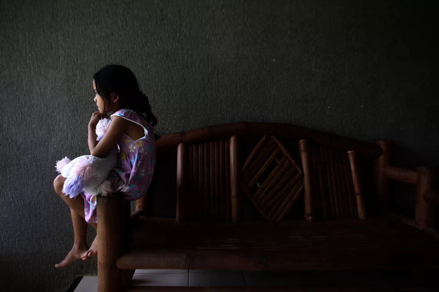 Ni&ntilde;a de perfil, sentada en el posabrazos de un sill&oacute;n, casi en la oscuridad, abrazando un peluche.